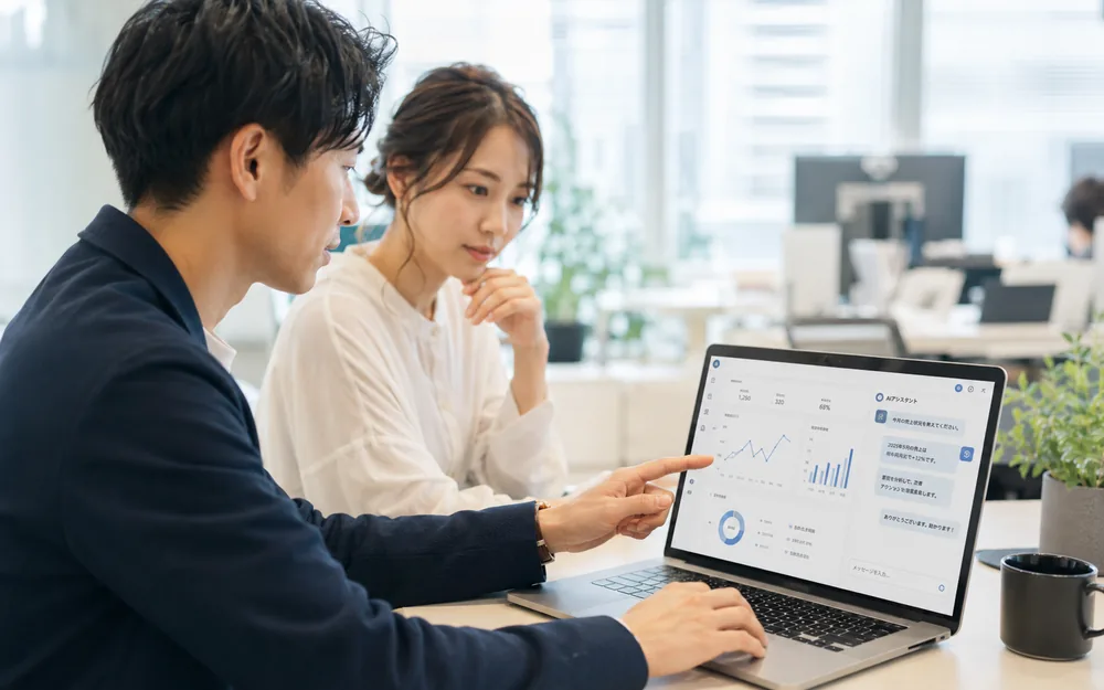 オフィスで社員2人がノートPCのダッシュボードとAIアシスタントの画面を見ながらデータを業務に活用している様子